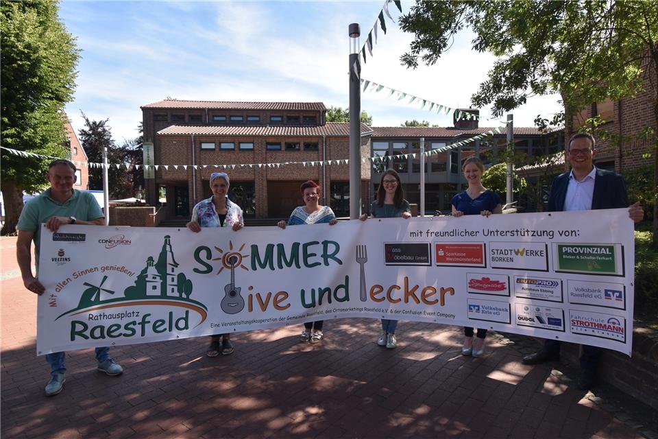 Bernhard Gelking von der „Confusion Event Company“ (links), Bürgermeister Andreas Grotendorst (rechts) und das Team vom Ortsmarketing Raesfeld präsentieren den Banner für die Veranstaltungsreihe „Raesfelder Sommer – Live und Lecker“ auf dem Rathausplatz. 
