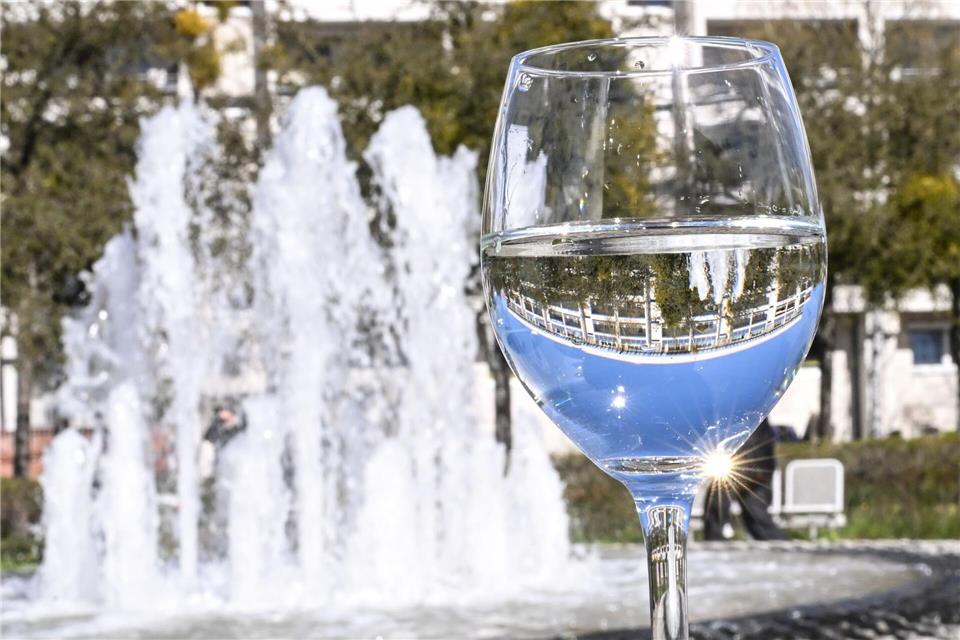 Der Brunnen auf dem Clara-Zetkin-Platz spiegelt sich in einem mit Wasser gefüllten Weinglas.Britta Pedersen/dpa