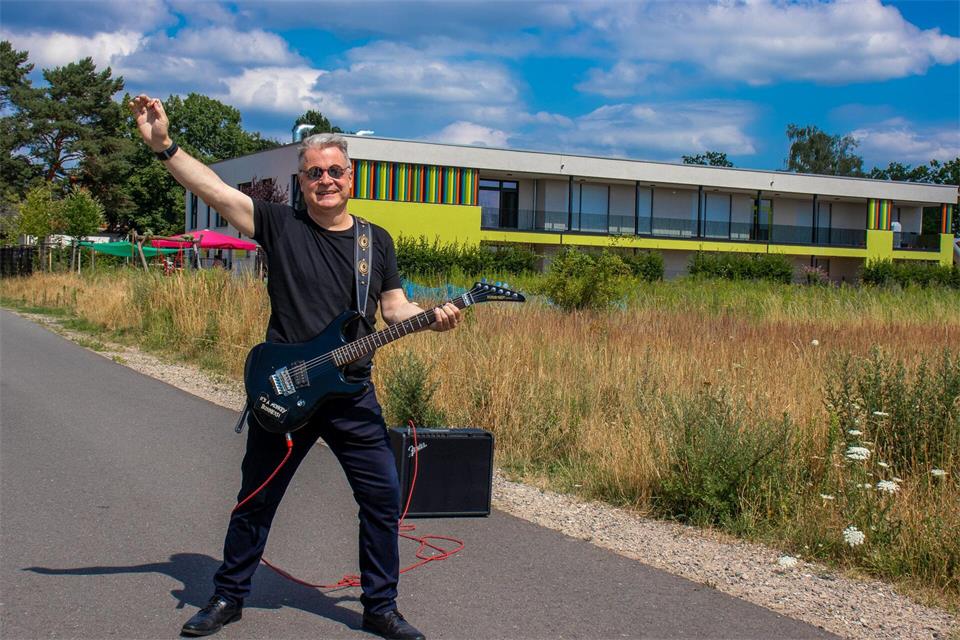 Der Bürgermeister von Stahnsdorf steht mit E-Gitarre vor einer Kita. Die Straßen im neuen Wohngebiet der Gemeinde sollen künftig die Namen von Rockmusikerinnen und -musikern tragen.-/Gemeinde Stahnsdorf/dpa