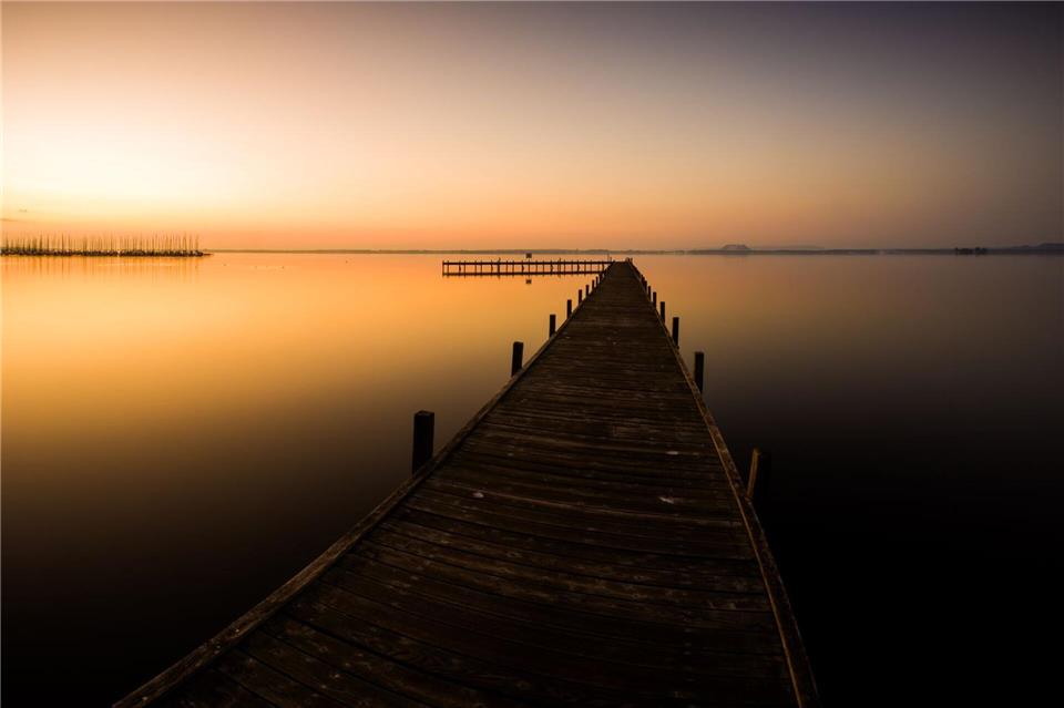 Der Himmel über einem Steg am Steinhuder Meer in der Region Hannover ist bei Sonnenaufgang orange.Moritz Frankenberg/dpa