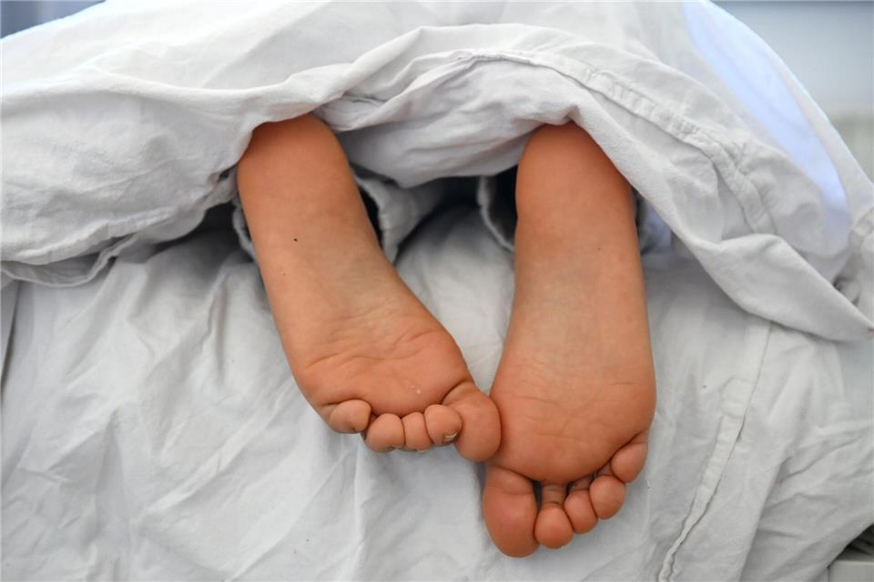 Die nackten Füße und Pyjamahose einer Frau sind unter einer Bettdecke zu sehen.Elisa Schu/dpa