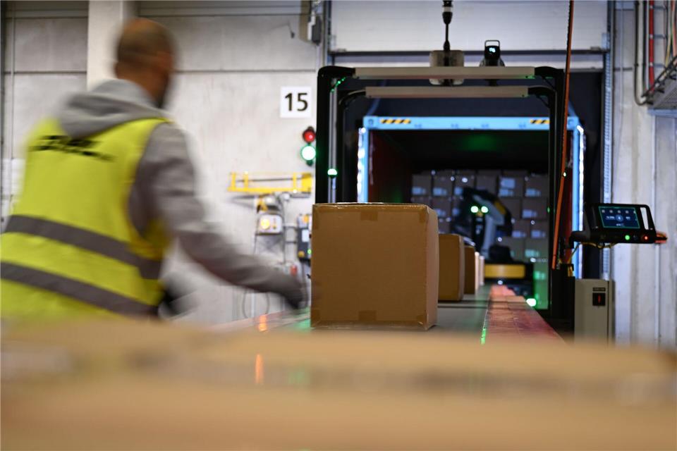 Ein DHL-Mitarbeiter nimmt Pakete in Emfpang - im Hintergrund ist ein Roboter zu sehen, der die Pakete aus einem Container nimmt und sie auf ein Förderband legt. (Archivbild)Wolf von Dewitz/dpa