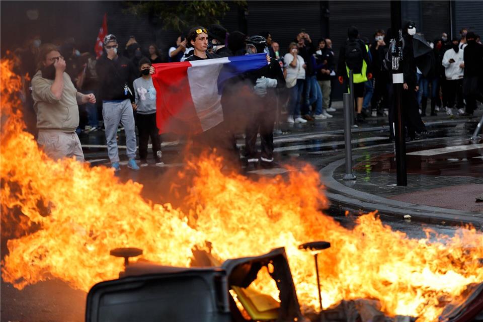 Ein Demonstrant der Protestbewegung „Bloquons Tout“ (Alles blockieren) zeigt eine französische Nationalflagge neben brennenden Mülltonnen.Jean-Francois Badias/AP/dpa