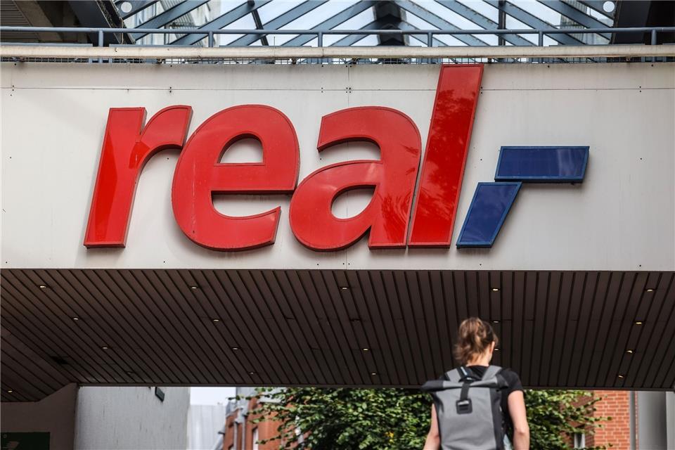 Ein Logo hängt über dem Eingang eines Real Marktes in Köln.