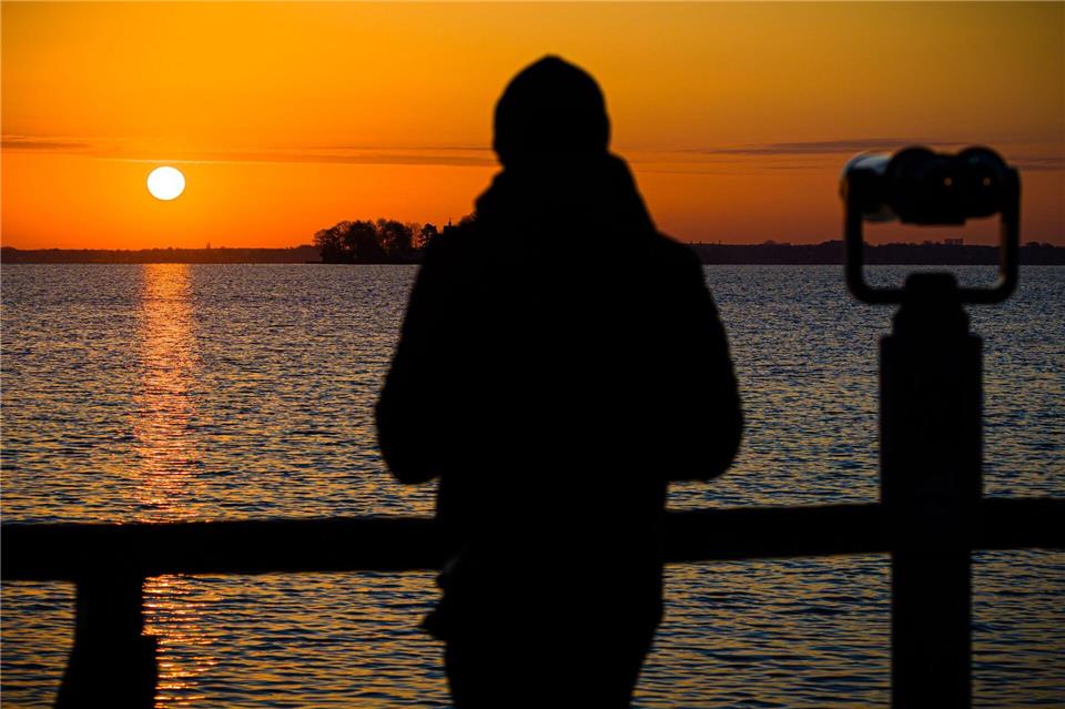 Ein Mann steht im Licht der aufgehenden Sonne neben einem Fernglas an einem Aussichtspunkt am Steinhuder Meer in der Region Hannover.Moritz Frankenberg/dpa