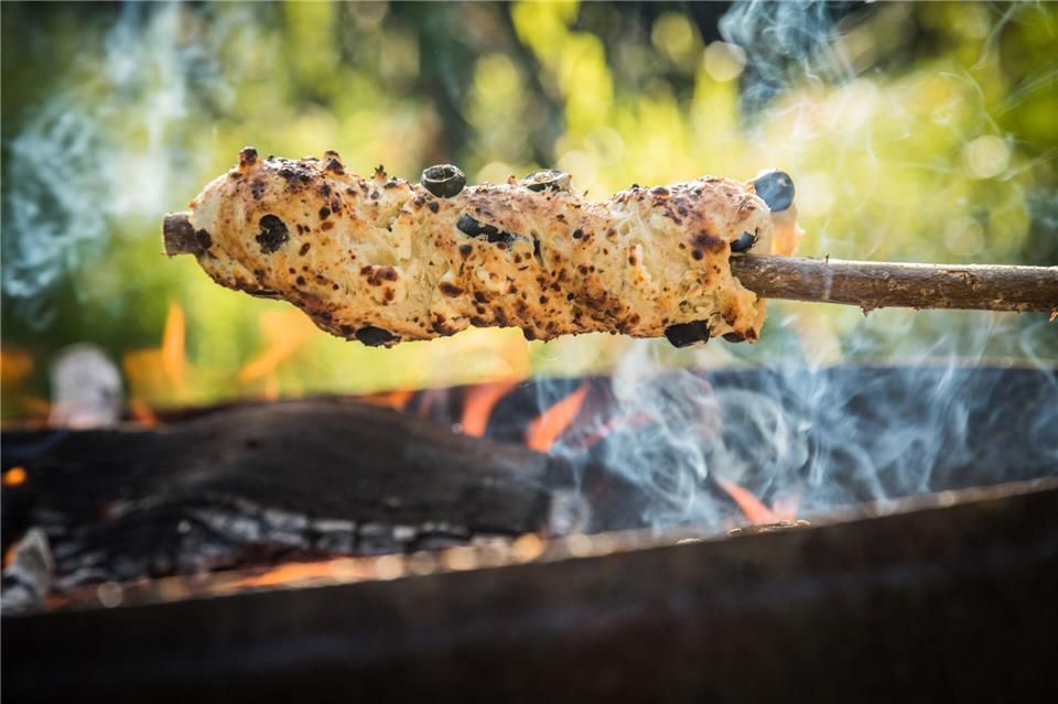 Ein Oliven-Feta-Knoblauch-Hefeteig ist um einen Haselnuss-Stock gewickelt und wird über dem Lagerfeuer gebacken.Christin Klose/dpa-tmn