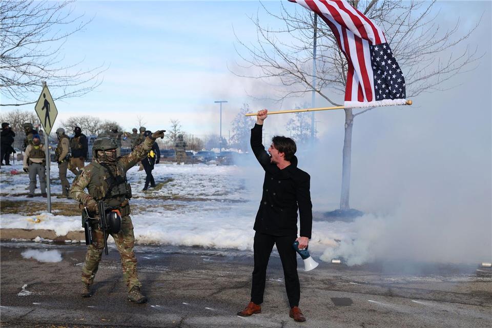 Ein Protestierender mit einer amerikanischen Flagge steht einem ICE-Beamten gegenüber.Adam Bettcher/AP/dpa