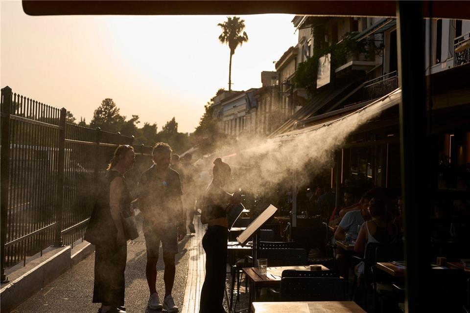Ein Touristenpaar geht an einer Sprühanlage vorbei, während ein Restaurantmitarbeiter sie im Athener Stadtteil Monastiraki zu ihrem Tisch führt.Petros Giannakouris/AP/dpa
