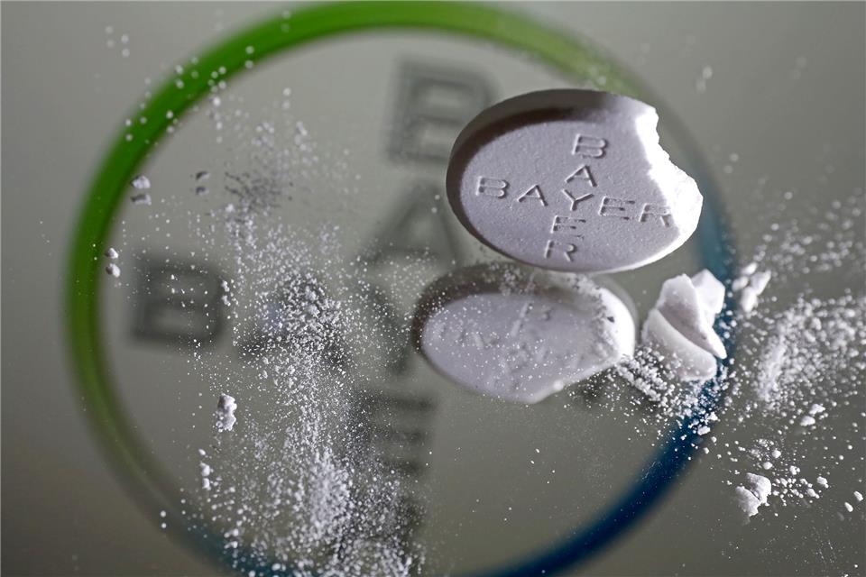 Eine Aspirin Tablette, an der eine Ecke abgebrochen ist, steht vor einem Bayer Logo auf einem Spiegel.