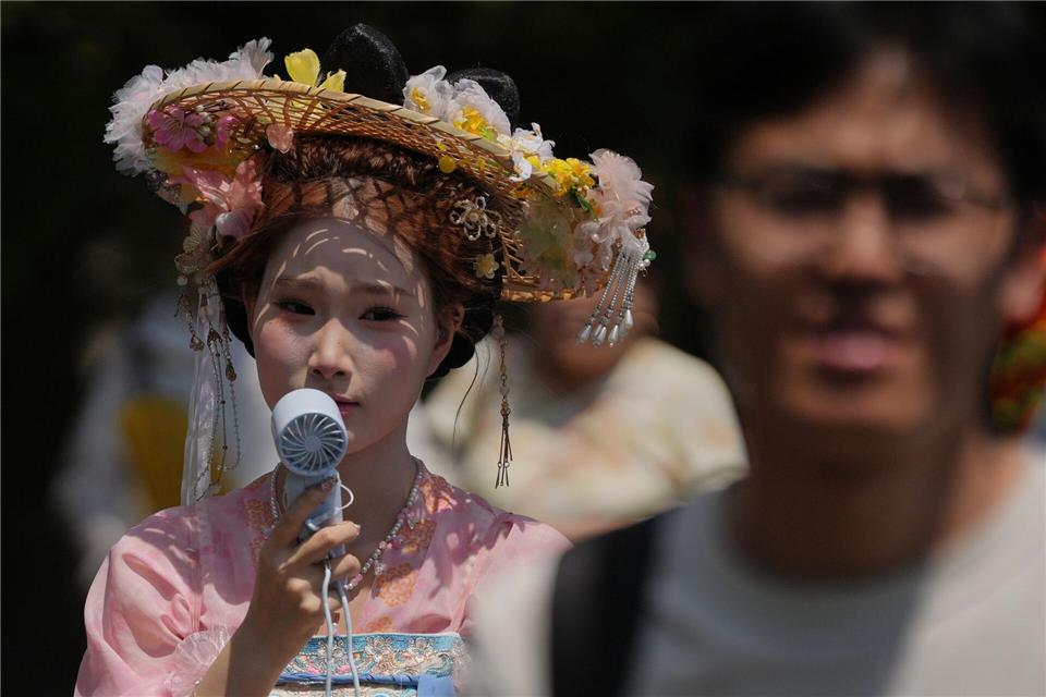 Eine Frau, die einen Hut mit Blumen und ein traditionelles Kostüm trägt, kühlt sich mit einem tragbaren elektrischen Ventilator ab, während Menschen an einem heißen Tag die Verbotene Stadt in Peking besuchen.Andy Wong/AP/dpa