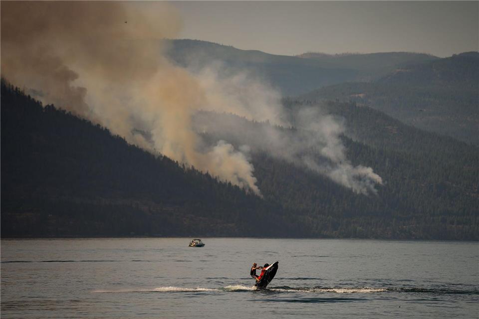 Eine Person fährt mit einem Jetski, währen im Hintergrund ein Wald brennt.DARRYL DYCK/The Canadian Press/dpa