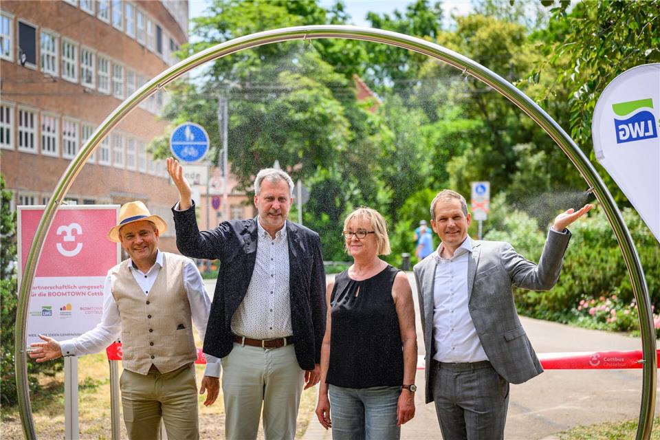 Erfrischender Wassernebel an heißen Tagen: Die Stadt Cottbus montierte ein Tor, das Wasser fein zerstäubt und für Abkühlung sorgen soll. Oberbürgermeister Tobias Schick (zweiter von links) hat es schon einmal getestet. Patrick Pleul/dpa