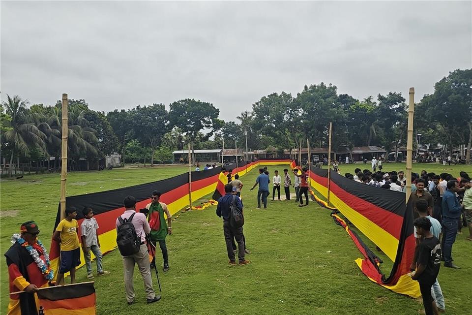 Fans haben sich an einer Seite versammelt, während eine riesige, 7,5 Kilometer lange deutsche Flagge auf dem Schulplatz von Nishchintapur im bangladeschischen Distrikt Magura entrollt wurde. Nazrul Islam/Nerob Hossain/dpa