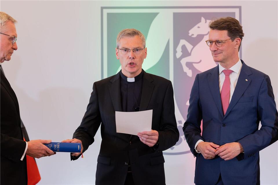 Heiner Wilmer (Mitte) legt bei seiner Vereidigung in der Staatskanzlei neben Ministerpräsident Hendrik Wüst (rechts, CDU), seine Hand auf eine Bibel.Rolf Vennenbernd/dpa