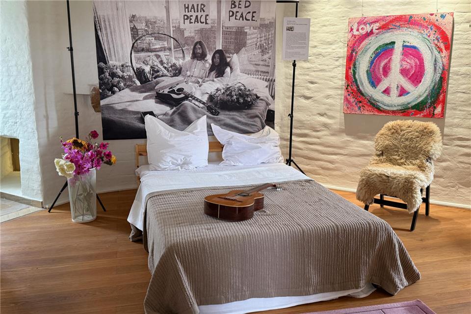 Im Zentrum der Installation steht ein einladendes Bett, über dessen Kopfende sich der Blick wie durch ein Fenster auf John Lennon und Yoko Ono während ihrer berühmten Bed-In-Aktion in Amsterdam richtet.