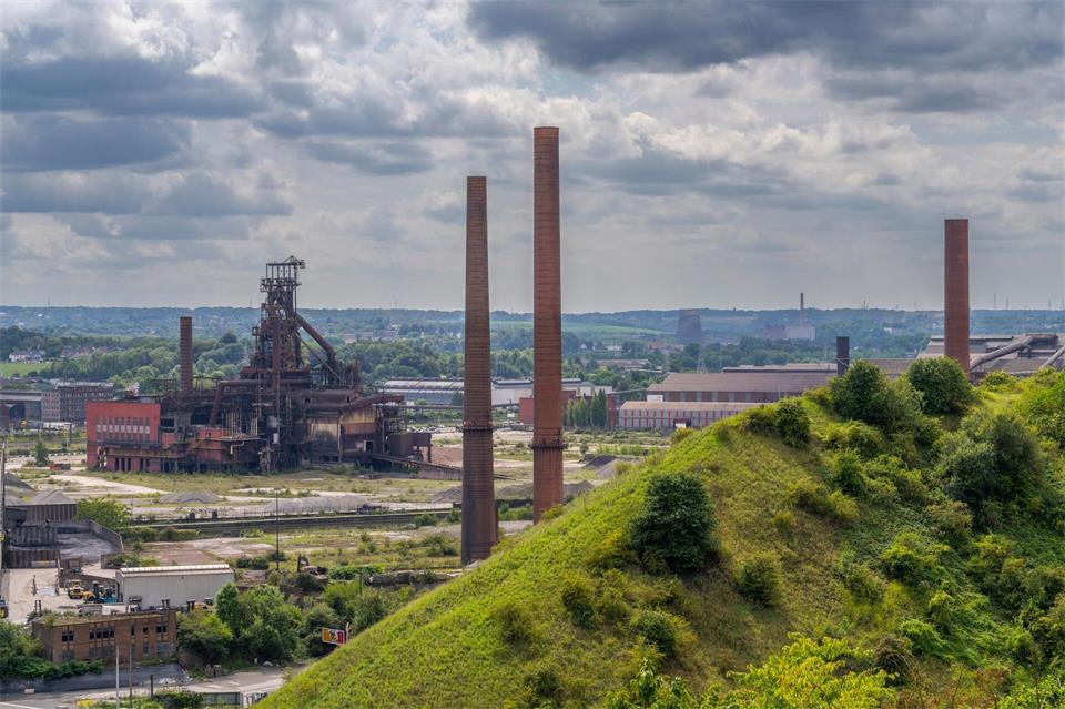 Industriegebiet von Charleroi: Blick über eine bewachsene Halde des Kohleabbaus am Wanderweg „Boucle Noire“, hinten Schornsteine und ein ehemaliger Hochofen eines Stahlwerks.Oliver Gerhard/dpa-tmn