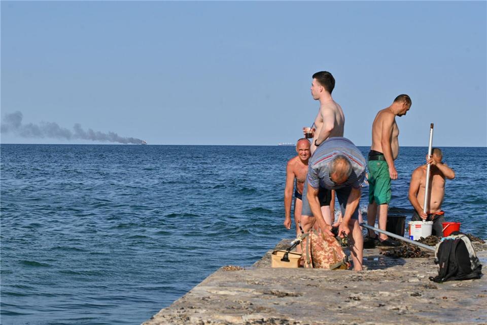 Menschen in Odessa sonnen sich, während am Horizont im Schwarzen Meer der brennende Getreidetransporter „Golden Leo“ zu sehen ist. (Archivbild)Michael Shtekel/AP/dpa