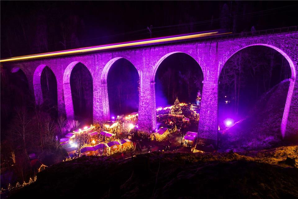 Menschen stehen auf dem Weihnachtsmarkt in der Ravennaschlucht, während über ihnen ein Zug über das Eisenbahn-Viadukt fährt. Philipp von Ditfurth/dpa