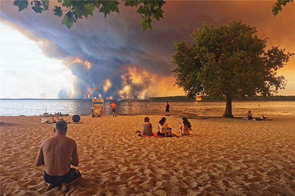 Strandbesucher sitzen am Strand von Moutchic in Lacanau im Südwesten Frankreichs, während Rauchwolken von einem Waldbrand in den Himmel aufsteigen.Maximilien Lamy/AFP/dpa