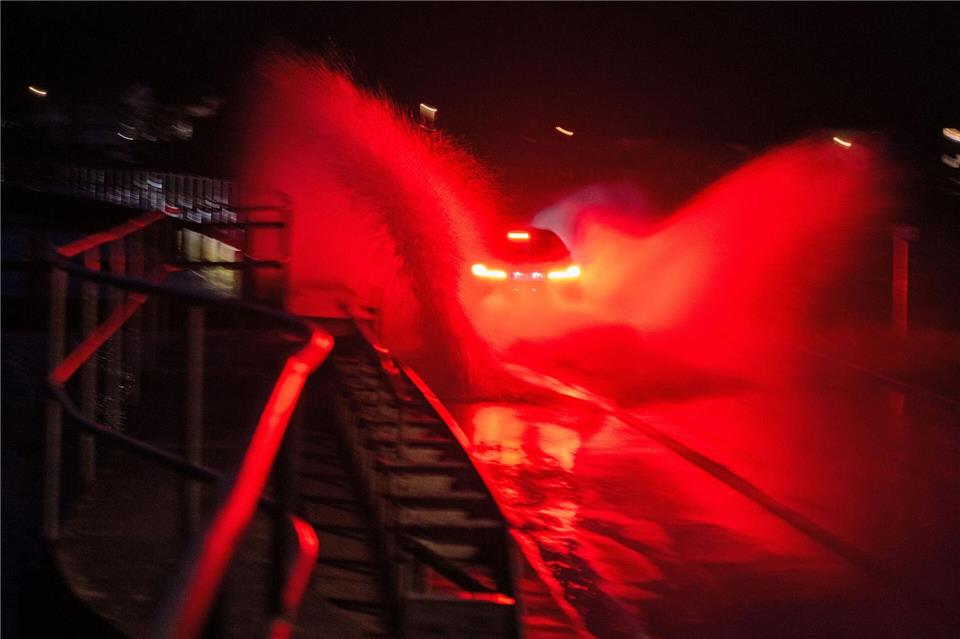 Wasser spritzt, während ein Auto über eine überschwemmte Straße zwischen Gleusdorf (Landkreis Haßberge, Unterfranken) und Busendorf (Landkreis Bamberg, Oberfranken) fährt.Pia Bayer/dpa