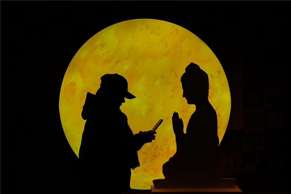 „Zwiegespräch“:  Eine Frau surft auf ihrem Smartphone vor einer Buddha-förmigen Pappfigur, die in einem Einkaufszentrum in Peking aufgestellt ist.Andy Wong/AP/dpa